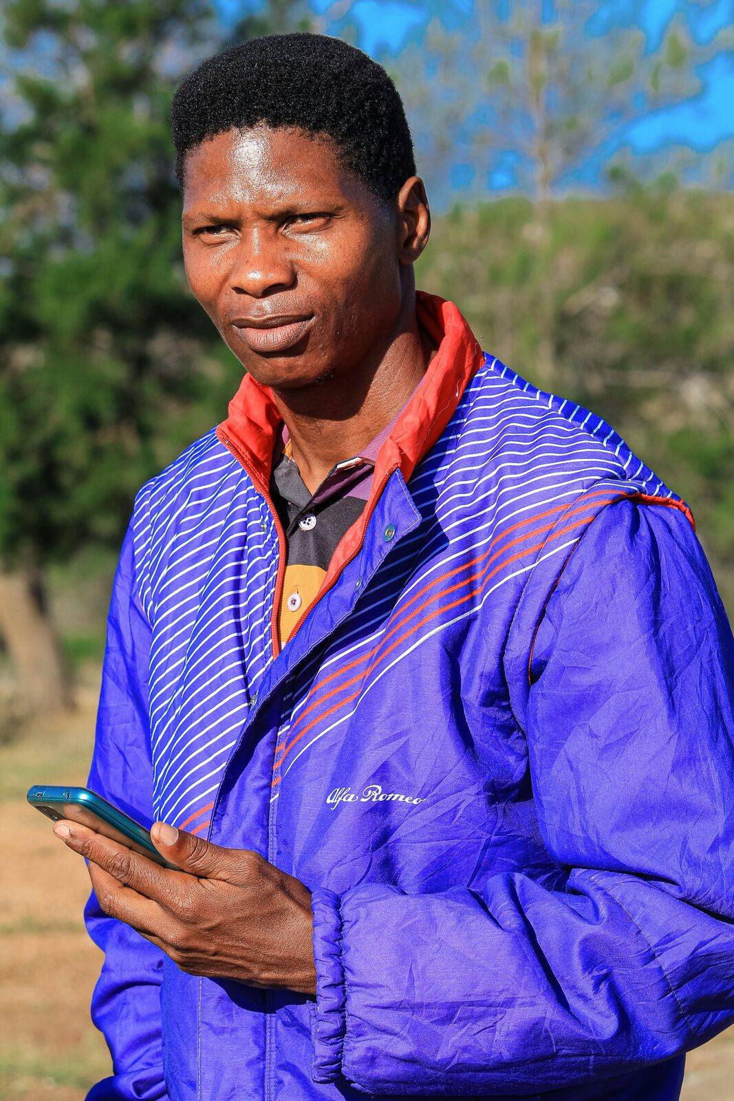 A man in a blue jacket standing outdoors, holding a smartphone and looking into the distance.