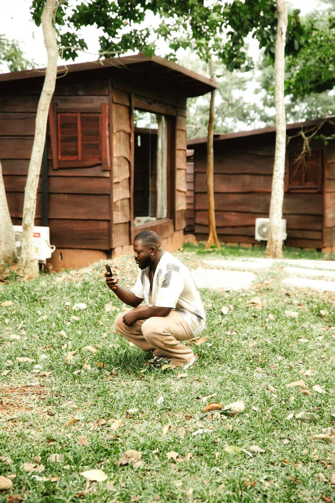 A man crouching on a lawn in front of a row of small wooden buildings, taking a photo with his phone.