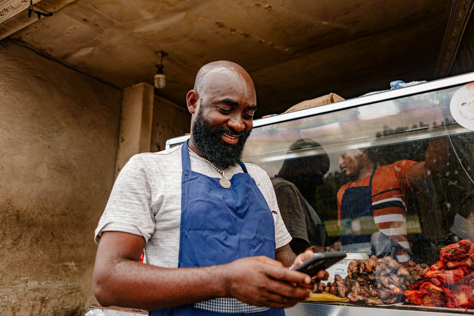 A smiling man in a blue apron checks his phone at the counter of a food stall.