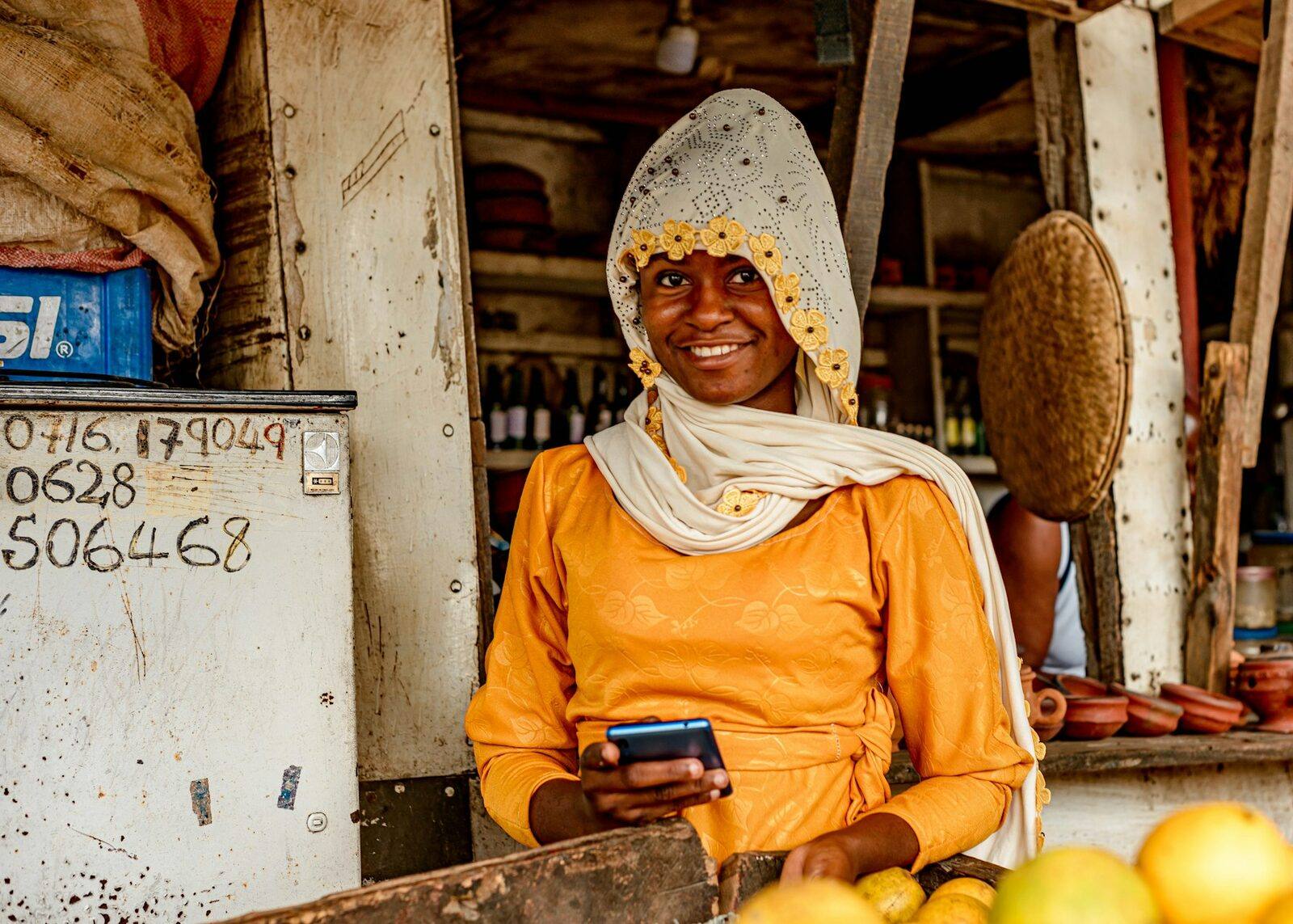 A smiling woman in an orange dress and headscarf holds a phone at her fruit stall.