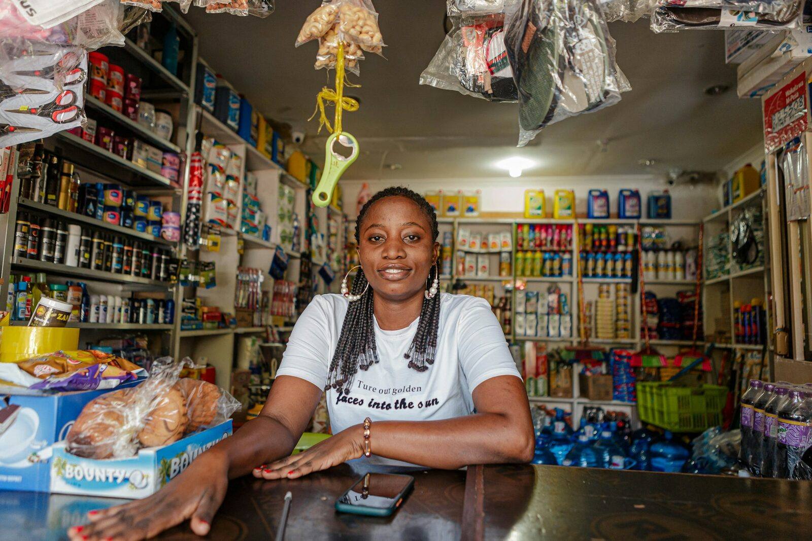A smiling shopkeeper leaning on the counter of a small general store stocked with groceries and household goods.